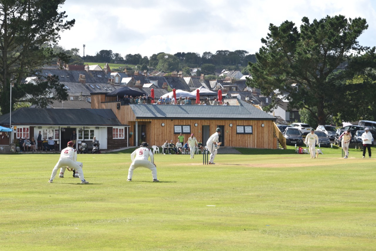 A Saturday match in progress at Egloshayle Park: fielders in whites, the pavilion terrace full, Wadebridge rising behind the trees.